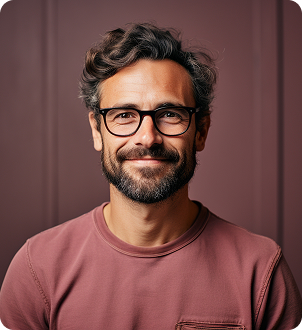 Smiling man with curly hair, beard, glasses, and a maroon shirt against a brown background.